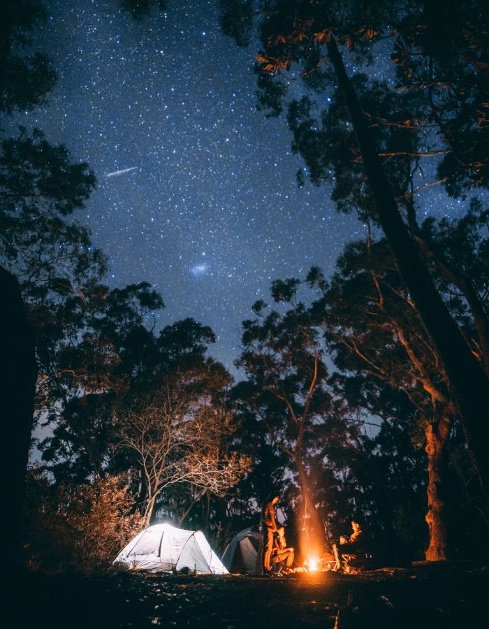 A tent pitched at a campsite with a dramatic starry night sky and three people sitting around a campfire
